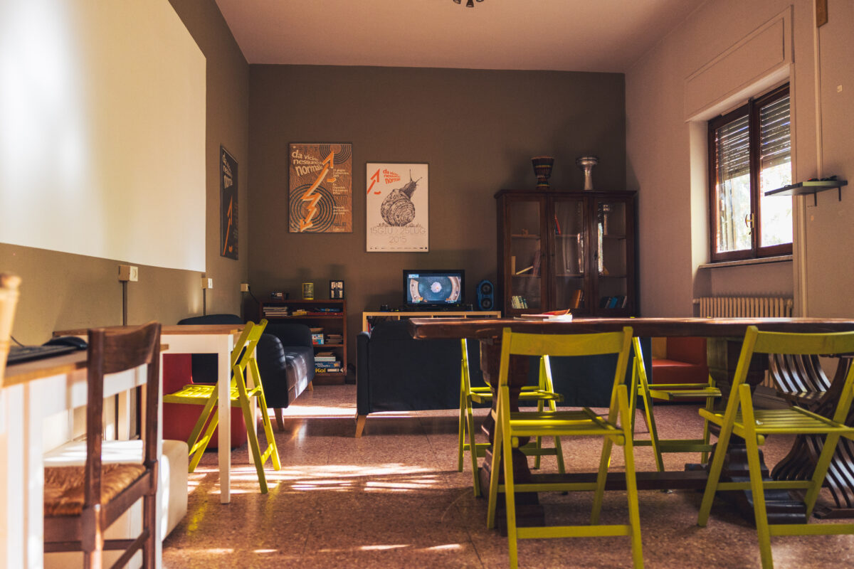 Sala comune luminosa con tavoli e sedie verdi, divani scuri, posters alle pareti, finestra ampia e mobile vetrina sullo sfondo con giochi e TV.