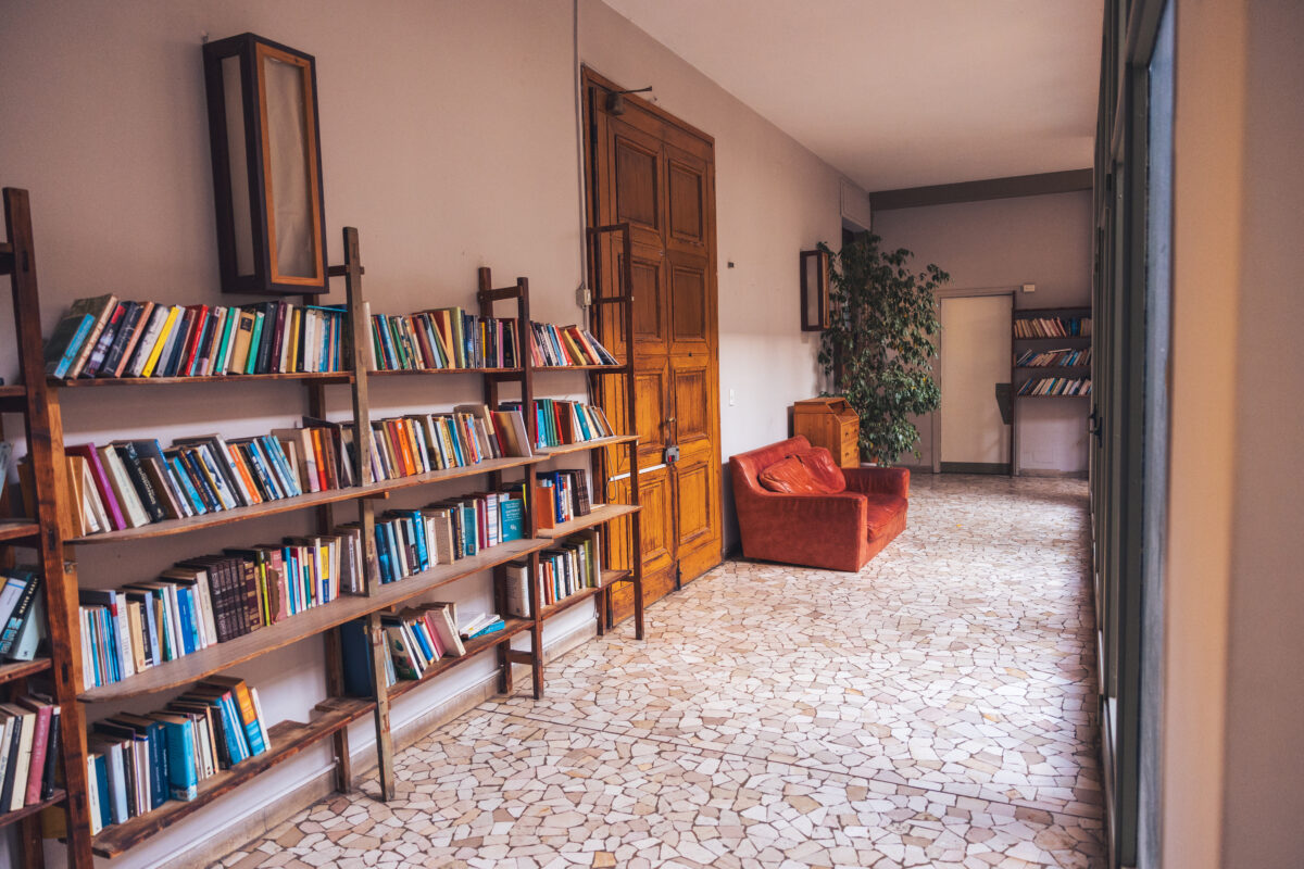Corridoio luminoso con librerie in legno piene di libri, grande porta in legno, poltrona rossa e pianta decorativa su pavimento in mosaico chiaro.