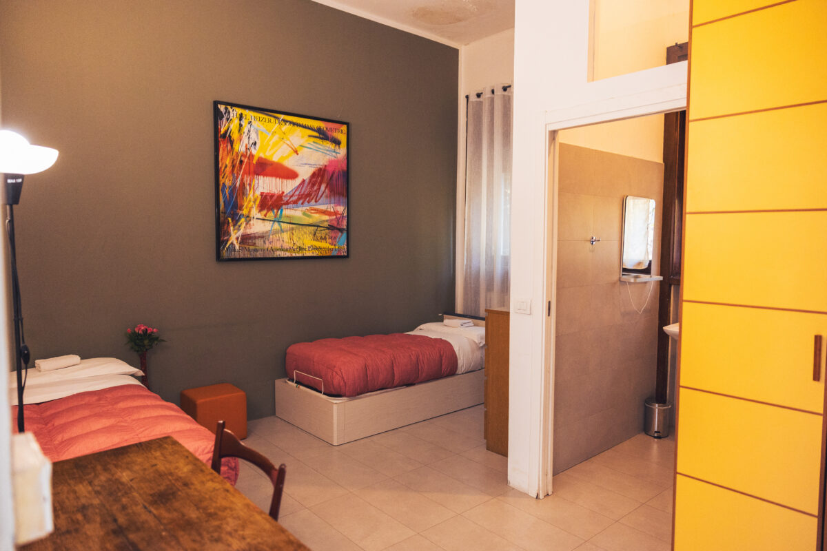 Camera con due letti singoli con coperte rosse, parete verde oliva con quadro colorato, scrivania in legno e ingresso al bagno con porta scorrevole bianca.