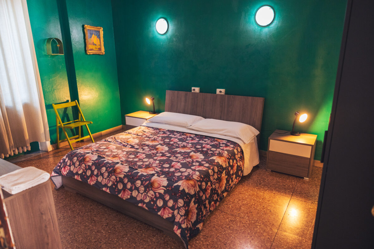 Camera da letto con pareti verde scuro, letto matrimoniale con coperta floreale, due comodini con lampade accese e una sedia gialla vicino alla finestra.