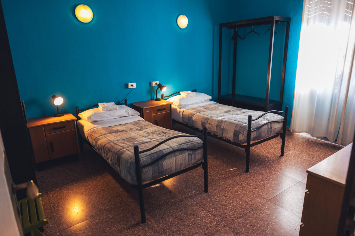 Camera con due letti singoli, pareti azzurre, comodini con lampade accese, appendiabiti aperto e finestra con tende bianche.