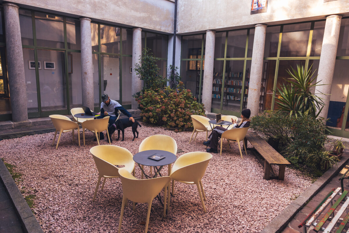 Cortile interno con tavoli e sedie gialle su ghiaia rosa, piante al centro e persone sedute a lavorare al laptop vicino a colonne e grandi vetrate.