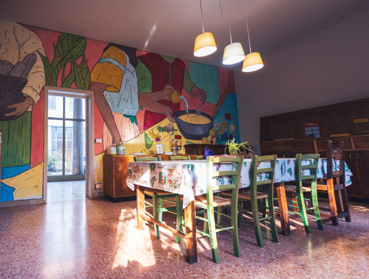 Sala da pranzo luminosa con tavolo lungo, sedie verdi, parete decorata da un grande murale colorato e lampade sospese.