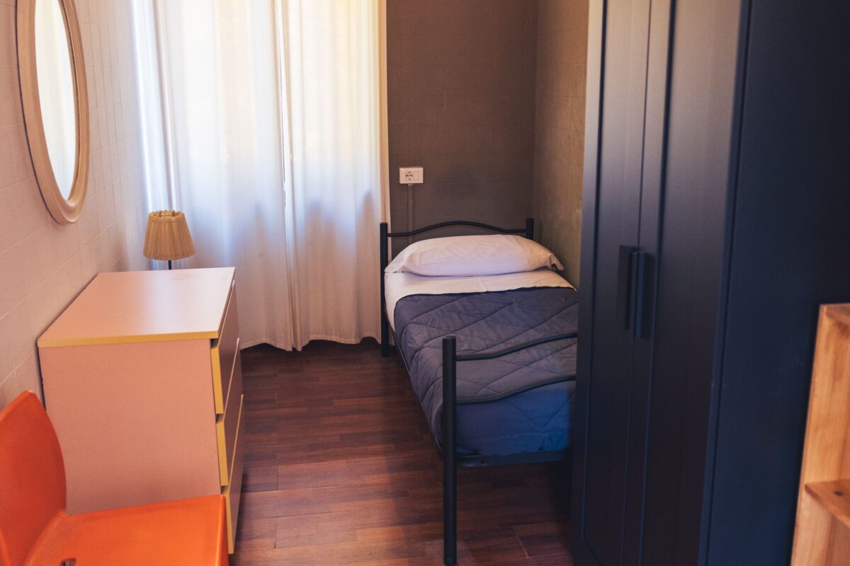 Camera singola semplice con letto singolo, comodino con lampada, cassettiera rosa, armadio scuro e finestra con tende bianche.