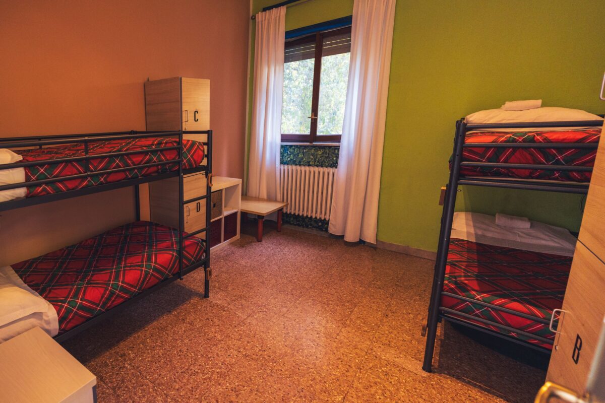 Camera semplice con due letti a castello, biancheria a quadri rossi, armadietti in legno, finestra con tende bianche e pareti colorate.